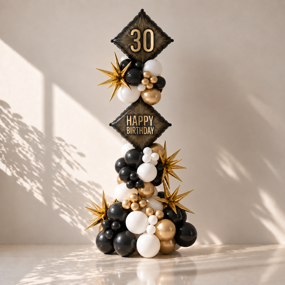 A 30th birthday balloon decoration tower featuring black, white, and gold balloons, starburst accents, and diamond-shaped signs reading '30' and 'HAPPY BIRTHDAY', set against a warm beige wall with soft window shadows.