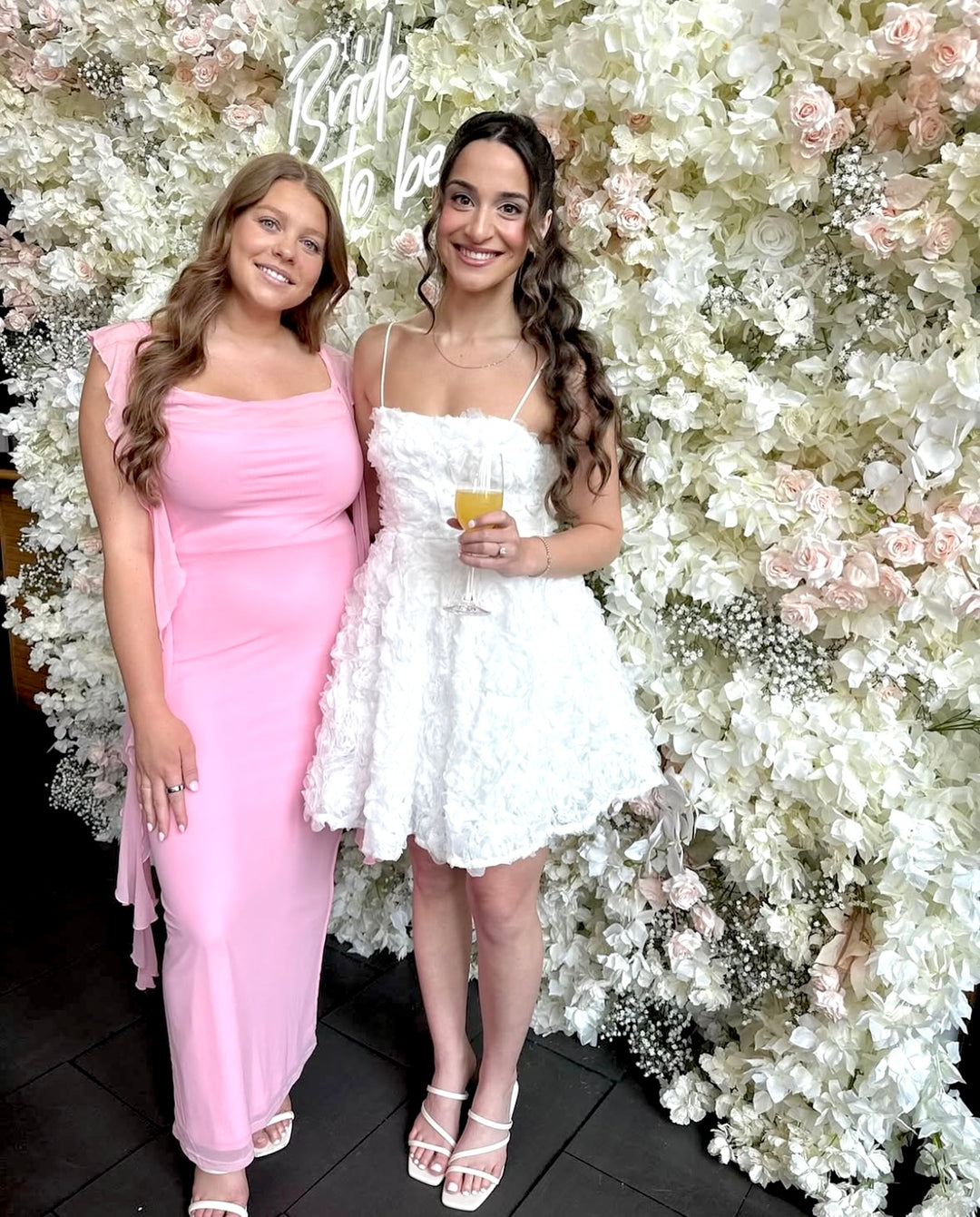 Two women posing in front of a floral backdrop with 'Bride to be' text.