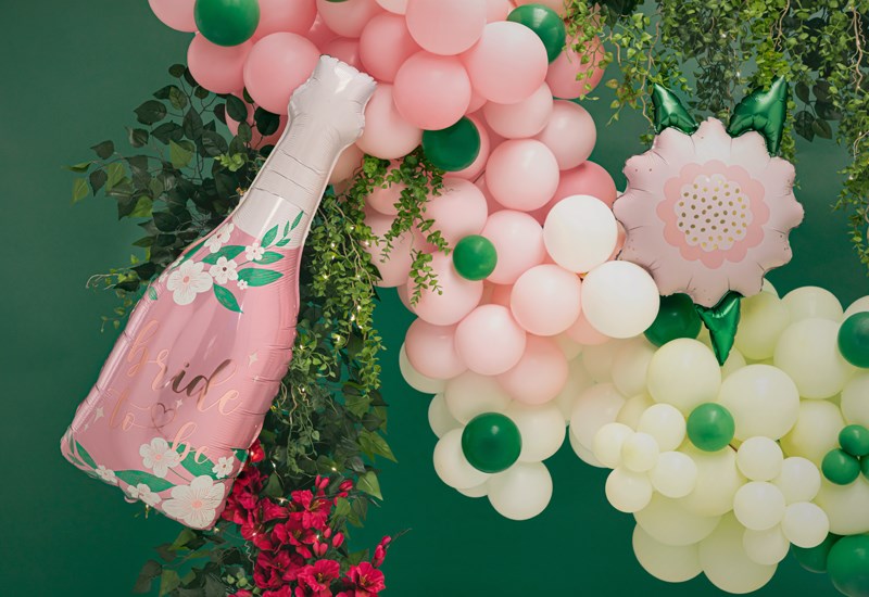 Decorative arch made of balloons and flowers with a pink champagne bottle balloon on a green background.