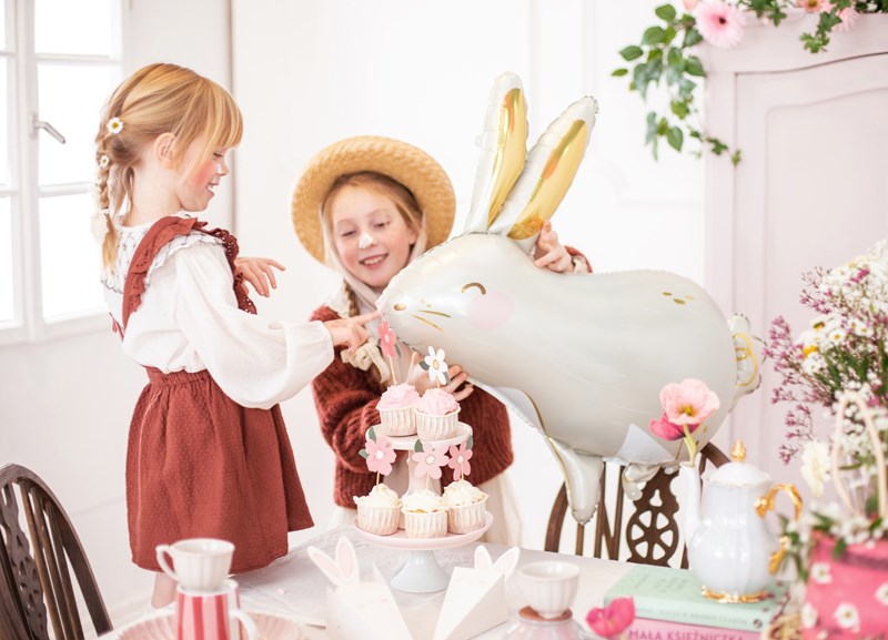 Two children at a tea party with a large plush rabbit, flowers, and a teapot.