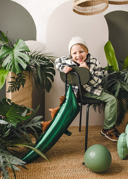 Child sitting on a chair with a green Dinosaur tail balloon  in a room with plants.