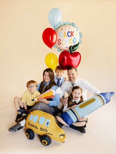 Family with children holding back-to-school items including balloons, an apple, a school bus, and a crayon.