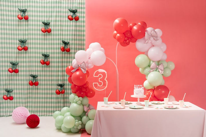 Decorative setup with balloons and a cake on a table against a pink wall with cherry decorations.