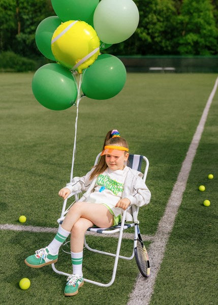 Child sitting on a chair with tennis-themed balloons and balls on a grass court.