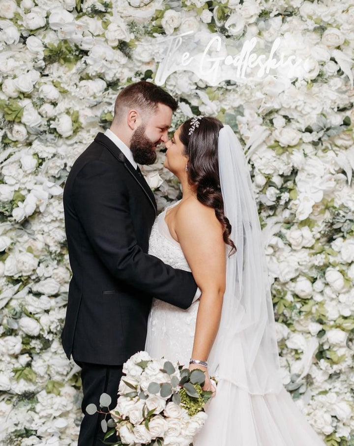 Wedding couple embracing in front of a white floral wall.