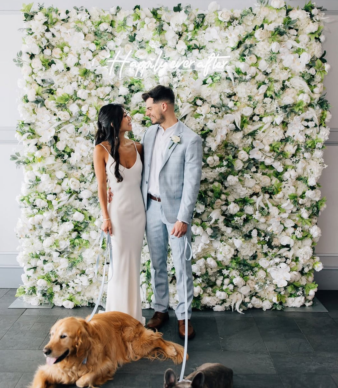 Couple standing in front of a floral wall with a dog at their feet.