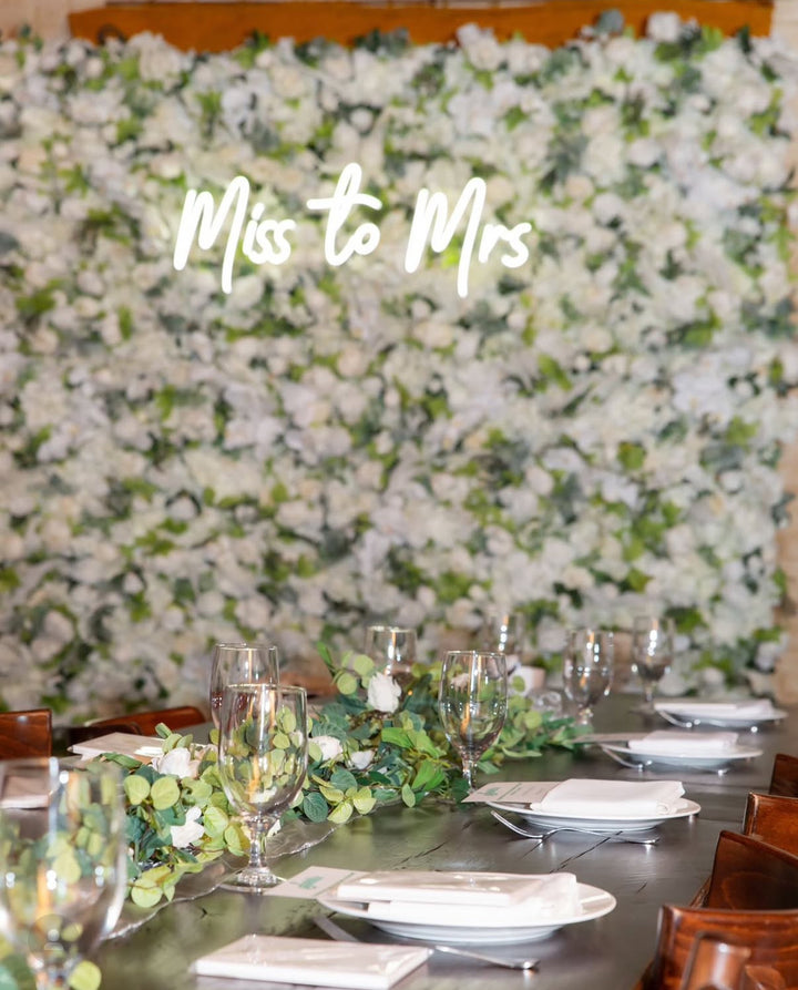 Decorated table with floral arrangements against a floral wall with 'Miss to Mrs' text.