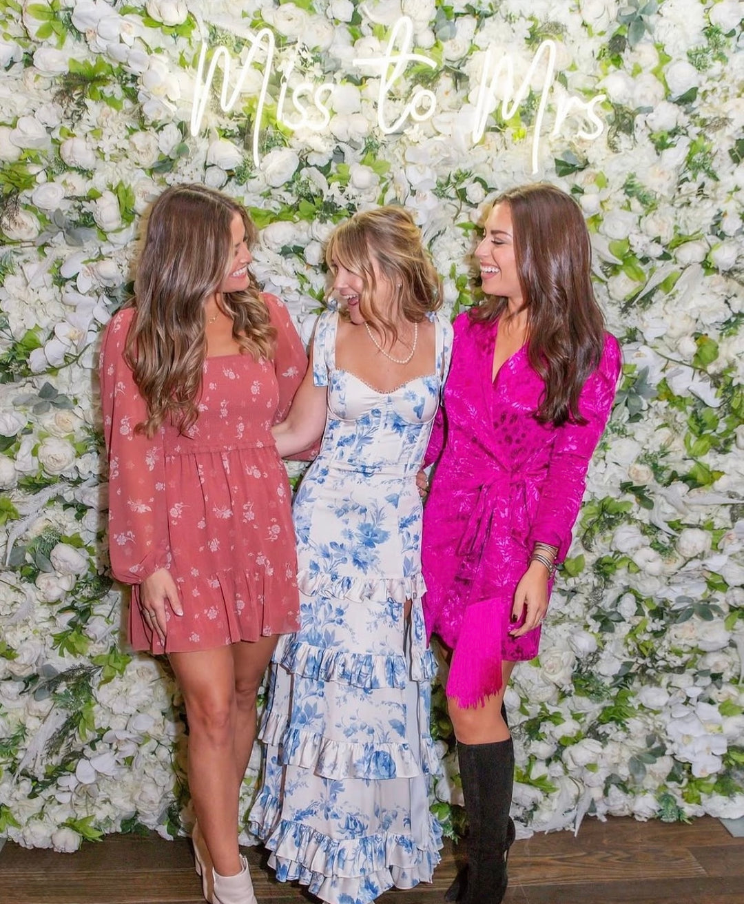 Three women posing together in front of a floral wall with 'Miss Dior' sign.