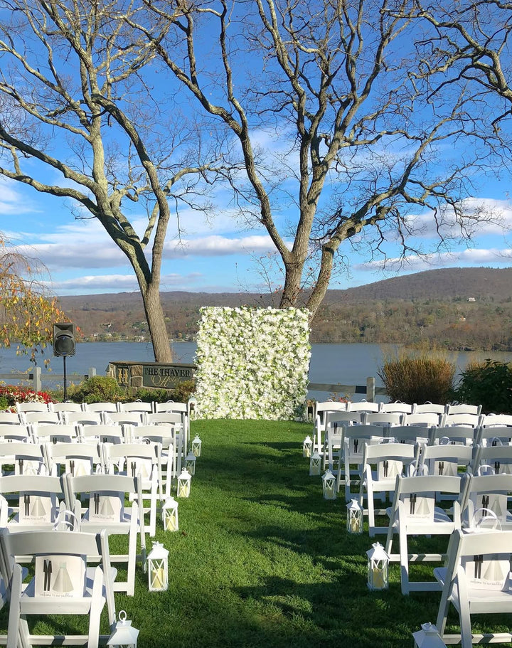 Outdoor wedding setup with white chairs and floral backdrop by a lake.