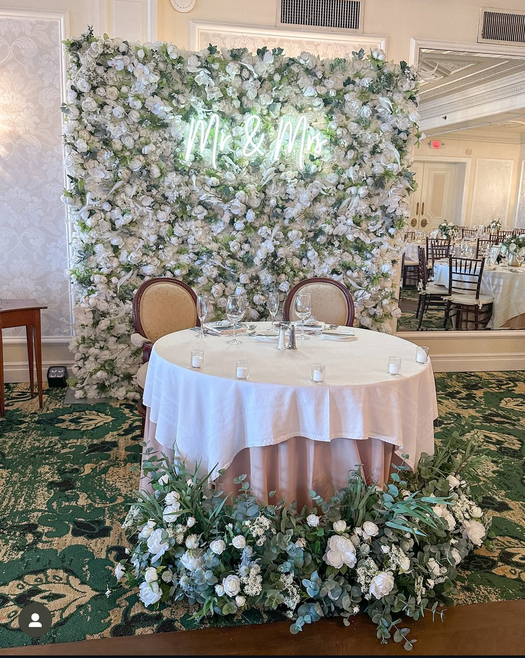 Decorated wedding table with floral backdrop and 'Mr & Mrs' sign in a banquet hall.