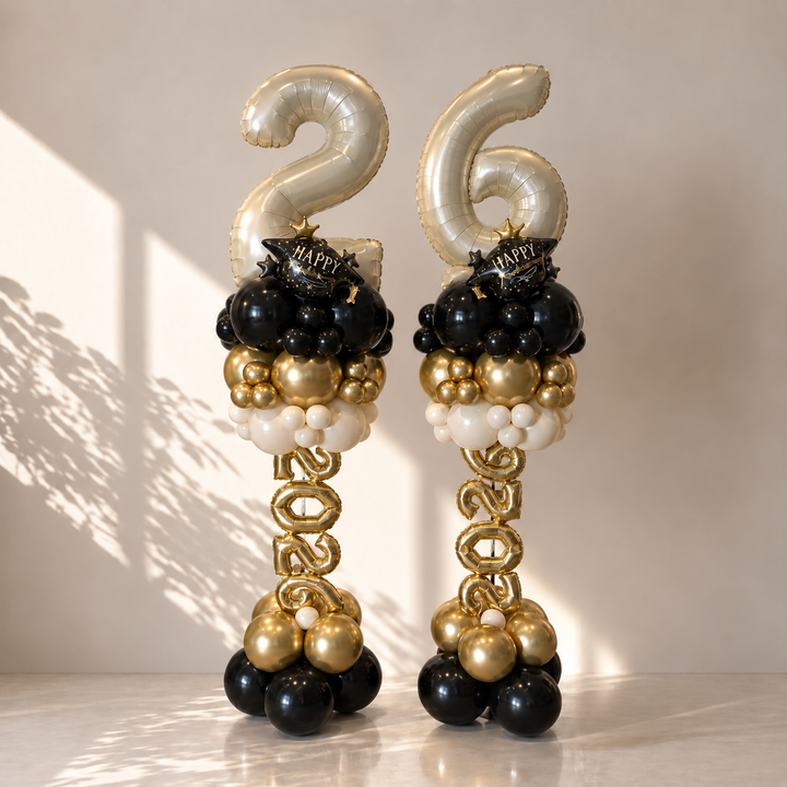 Two balloon sculptures shaped like pillars, decorated with large metallic gold foil balloons spelling '26' at the top, black and gold balloon clusters, white balloon accents, and gold 'HAPPY' balloons, set against a warm beige wall with soft shadows.