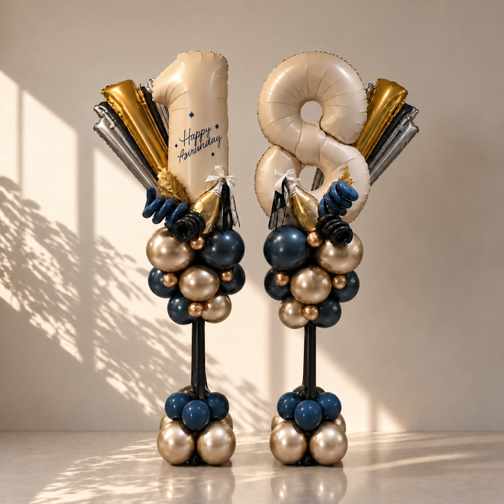 Two birthday balloon towers standing on a glossy floor against a warm beige wall. Each tower features large metallic foil balloons shaped like the numbers 8 and 1, with a 'Happy Birthday' message balloon, surrounded by clusters of navy blue, champagne gold, and silver balloons on black supports.