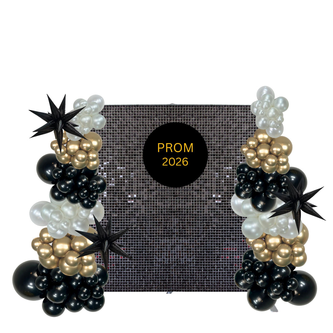 A black, white, and gold balloon garland set for a prom backdrop, featuring balloon clusters, star-shaped balloons, and a central black circle with gold text reading 'PROM 2026'.