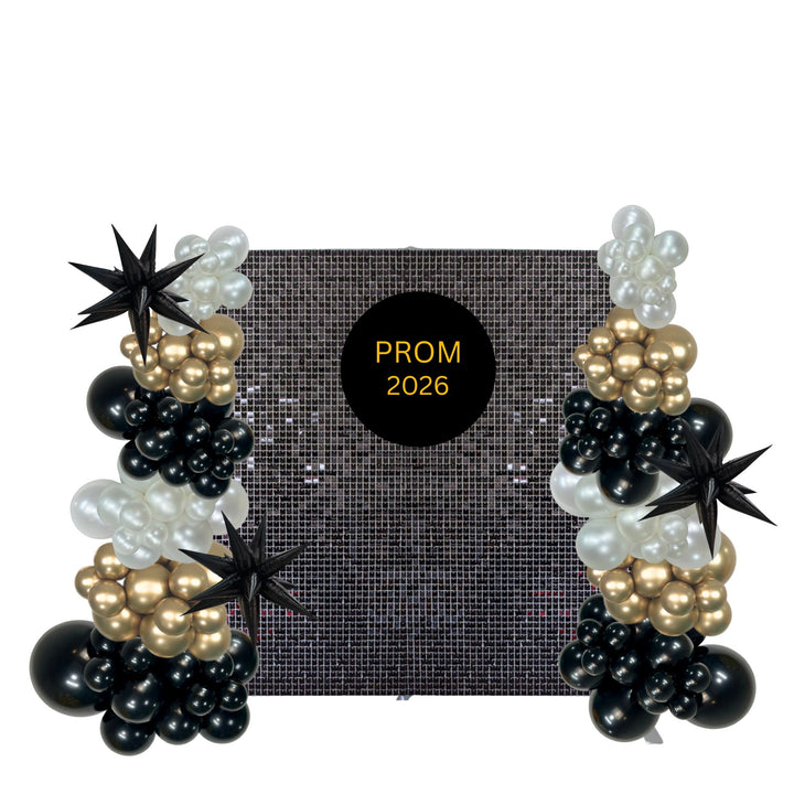 A black, white, and gold balloon garland set for a prom backdrop, featuring balloon clusters, star-shaped balloons, and a central black circle with gold text reading 'PROM 2026'.