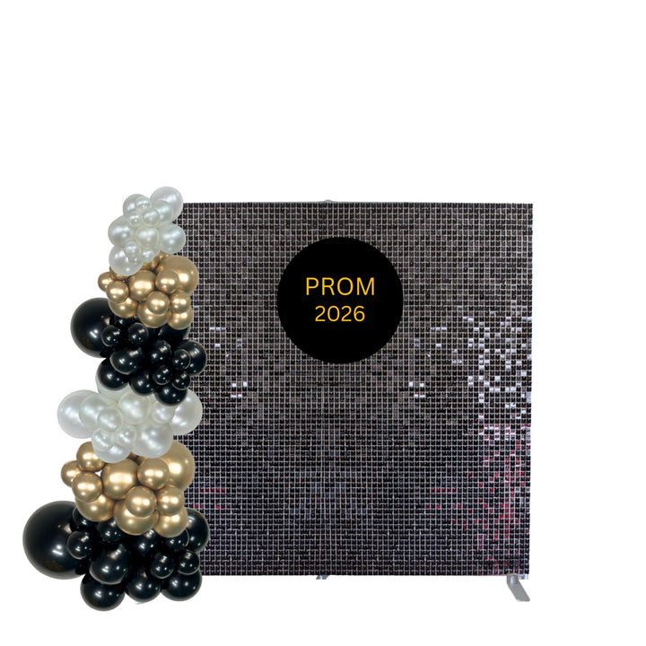 A black sequin prom backdrop with a black circular sign reading 'PROM 2026' in gold, styled with a balloon arch in black, white, and gold balloons on the left, against a plain white background.