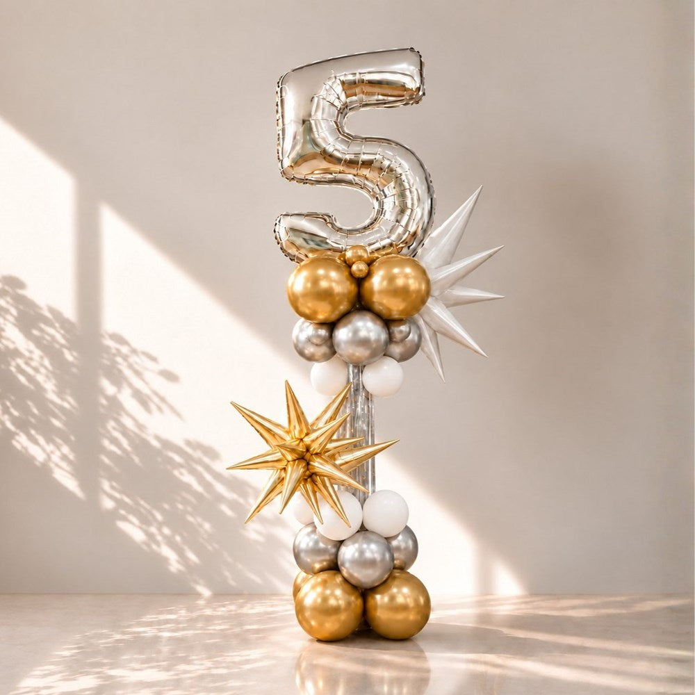A 5th birthday balloon decoration set featuring a large metallic silver number 5 balloon, gold and silver balloons, white balloons, and two metallic star-shaped balloons on a stand, set against a warm beige wall with soft window shadows.