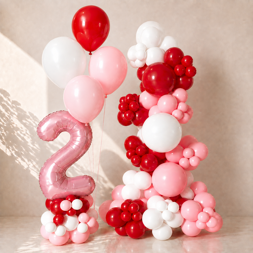 Pink, red, and white balloon decoration set including a large metallic pink foil balloon shaped like the number 2, standing balloons, and a cluster of round balloons in red, white, and pastel pink, arranged against a warm beige wall.