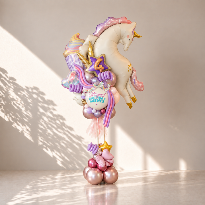 A whimsical unicorn-themed birthday balloon bouquet featuring a large white unicorn balloon with gold and pink accents, pastel purple and pink streamers, star balloons, and metallic rose-gold and pink balloons at the base, displayed indoors against a light gray wall with soft window shadows.
