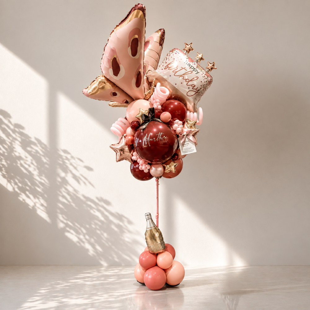 A decorative balloon bouquet stand with a cluster of pink, red, and peach balloons, foil stars, a gold bottle-shaped balloon, and a white balloon reading 'Happy Birthday,' displayed on a light neutral surface against a softly lit wall.