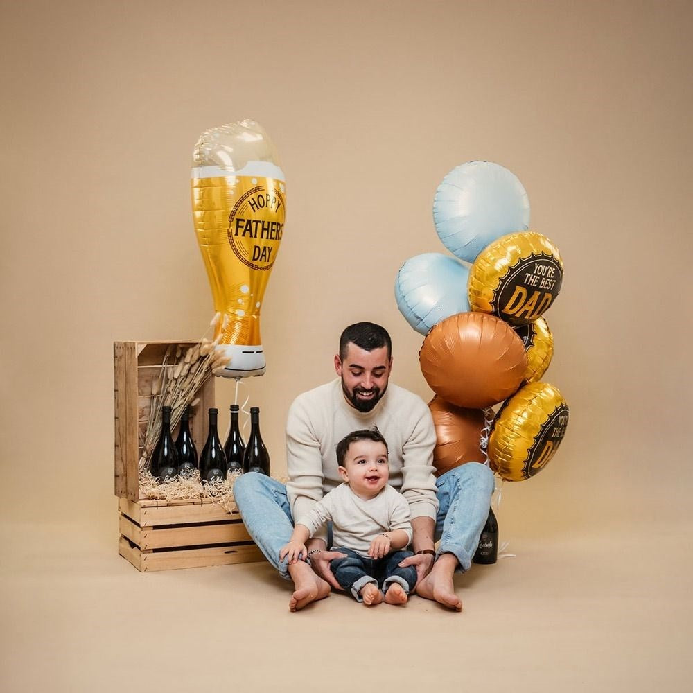 A father and child sitting together with Father's Day themed balloons and a wooden crate holding beer bottles, photographed in a studio setting against a warm beige backdrop.