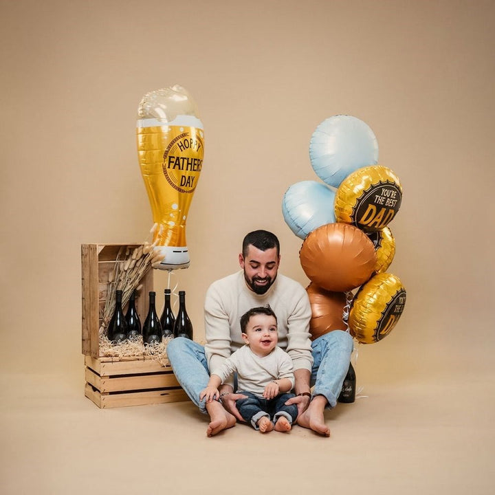 A father and child sitting together with Father's Day themed balloons and a wooden crate holding beer bottles, photographed in a studio setting against a warm beige backdrop.