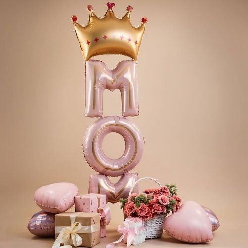 A pink and gold foil balloon decoration spelling 'MOM' with a large gold crown balloon on top, styled with pink heart balloons, a gift box, and a bouquet of flowers on a beige background.
