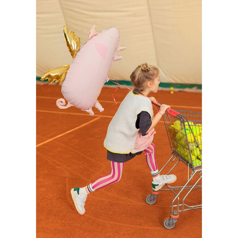A young girl in a vest and striped leggings running on a tennis court with a shopping cart full of tennis balls and a large pink flying pig balloon with gold wings.