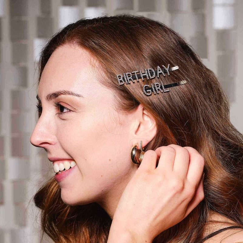 A smiling woman with brown hair wearing sparkly rhinestone "Birthday Girl" hair clips and gold hoop earrings.