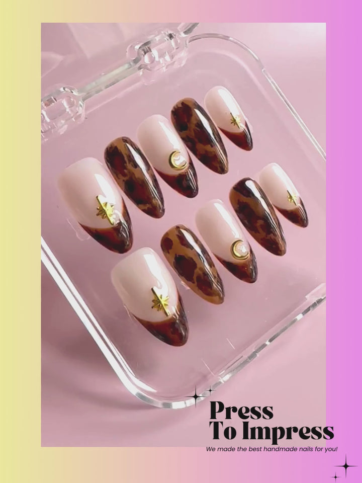 A Video shows A hand holds a clear plastic case containing press-on nails with a cream and brown leopard print design, gold crescent moon and star accents, and a gold leaf motif. The background is a soft pink-to-yellow gradient with the text 'Press To Impress' at the top.