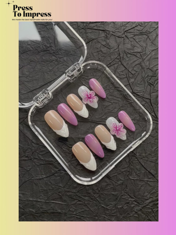 A Video shows A hand holds a clear plastic tray displaying false press-on nails. The nails feature gradient designs in pink and beige, French tips, and white floral accents.