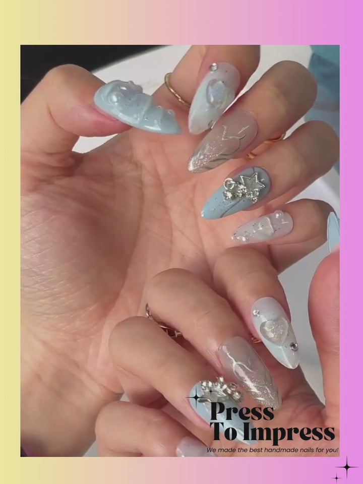 A video A hand holds a clear plastic case displaying ten decorative press-on nails. The nails feature light blue, white, and beige designs with glitter, pearl-like embellishments, and floral details.