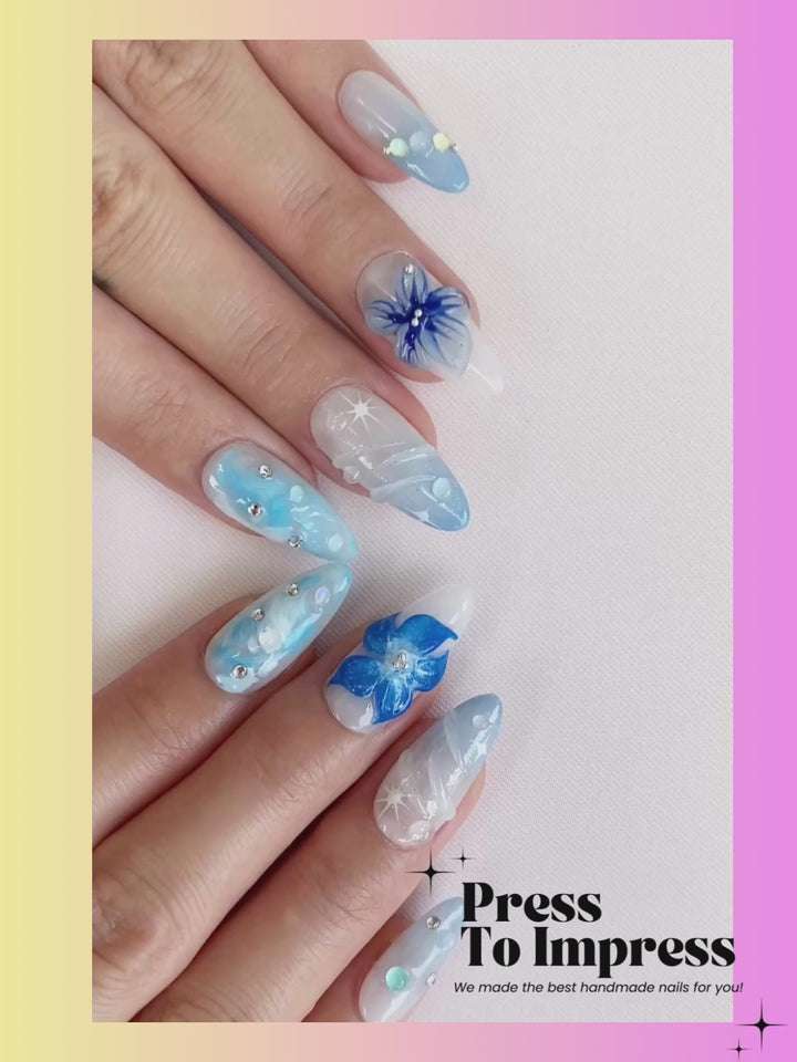 A Video shows A hand holds a clear plastic case displaying 10 press-on nails with blue and white floral, swirled, and glitter designs against a pink and yellow gradient background with 'Press To Impress' branding.