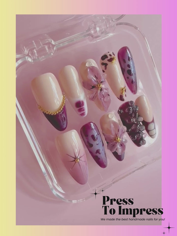 A Video shows A hand holds a clear plastic tray displaying press-on nails with a purple and beige floral/abstract design. The nails feature cream, purple, and dark plum tones with gold and pearl embellishments.
