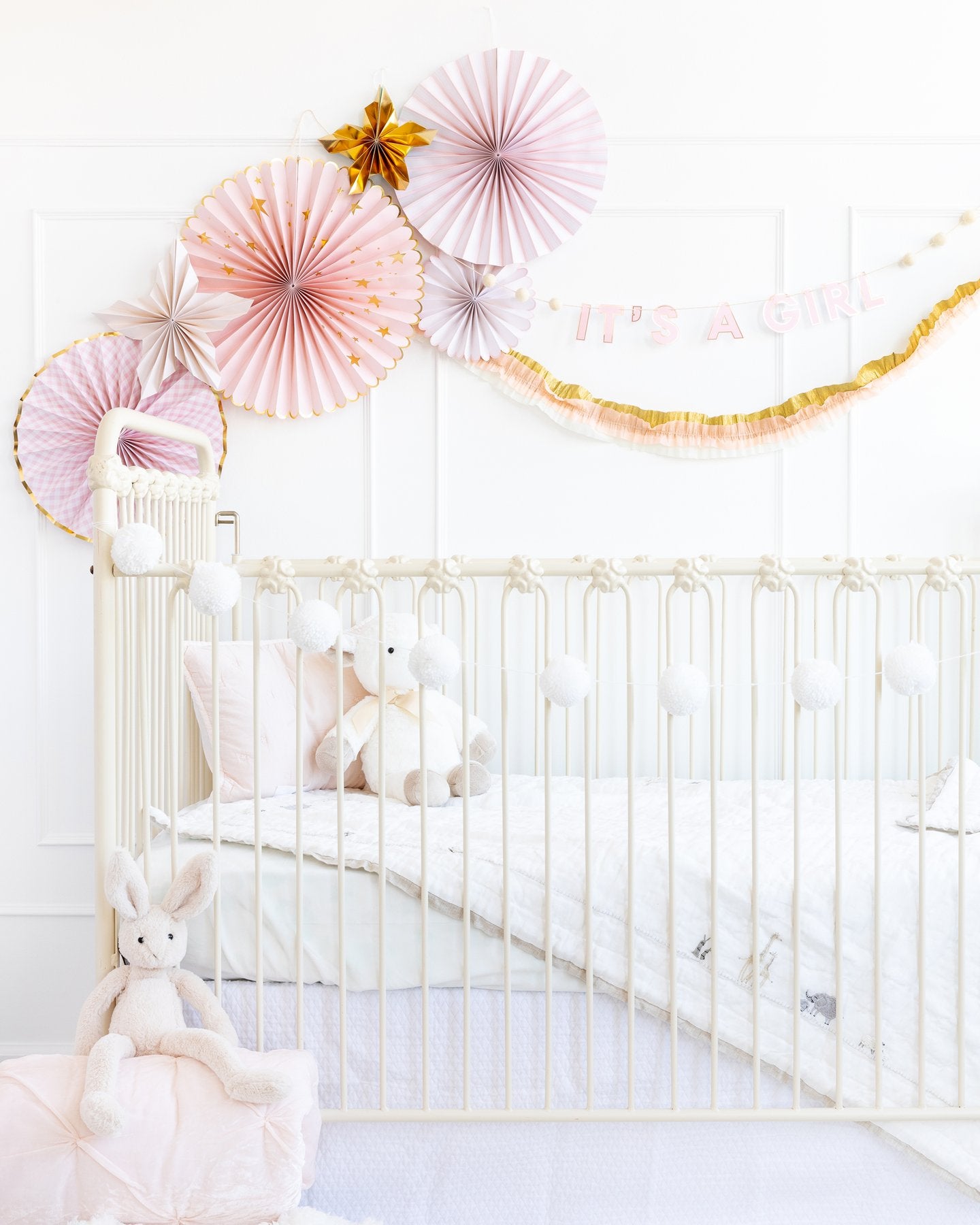 My Mind's Eye Baby Crea/Pink/Gold Crepe Paper Banner in package hanging above a white baby cot